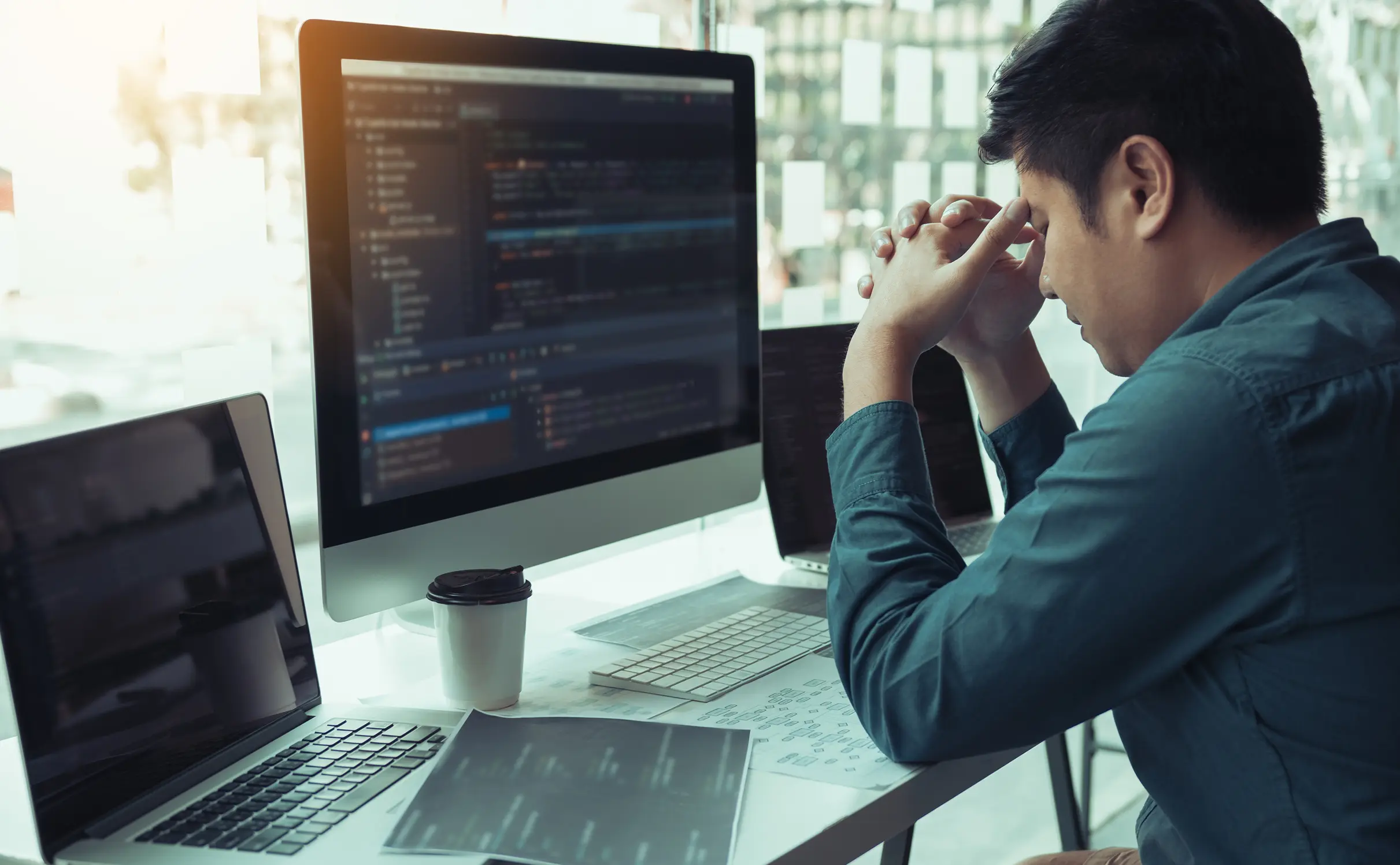 A stressed software developer sits at a desk with multiple monitors displaying code, holding his head in frustration, symbolizing slow and overwhelming product launch processes.