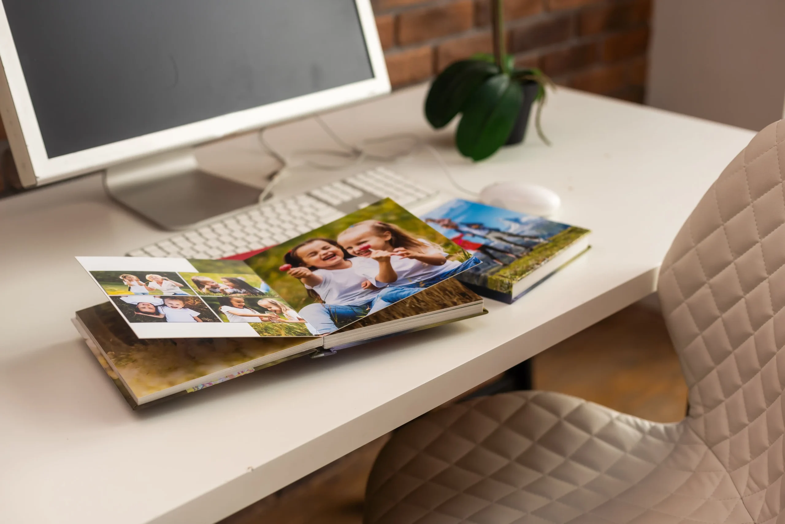 Open family photobook with colorful pages showing children, placed on a white desk with a keyboard and computer monitor